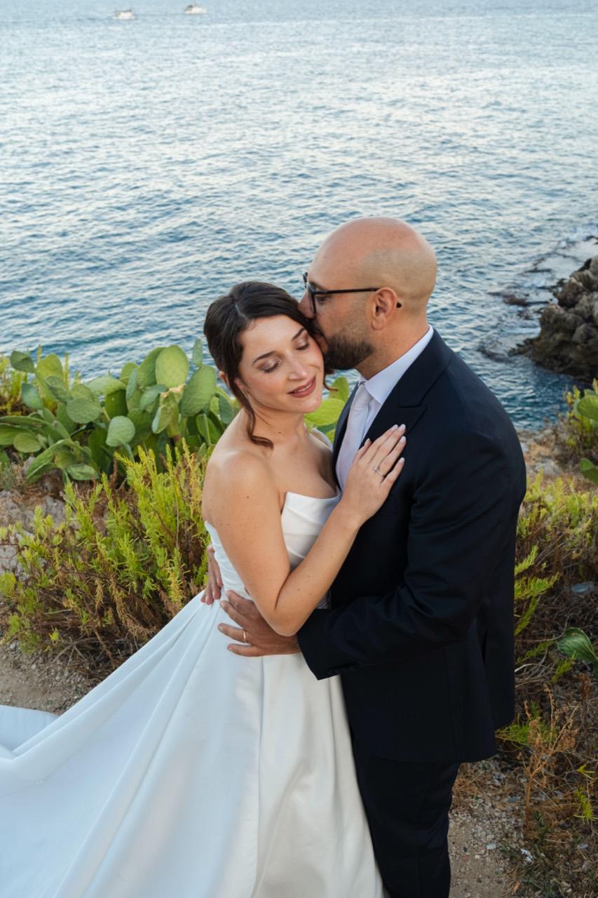 Lo sposo bacia la sposa sulla guancia davanti al mare.